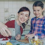 Smiling mother making selfie portrait together with young cute daughter cooking breakfast at home in kitchen. Family, cook, and people concept